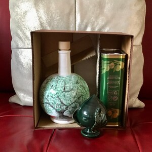 May include: A brown cardboard box with a green ceramic olive oil bottle, a green ceramic olive branch, and a green and gold can of olive oil. The box is open and the items are visible inside.