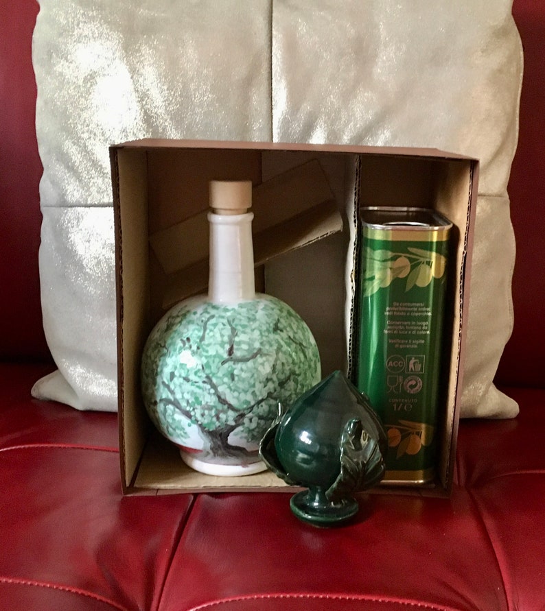 May include: A brown cardboard box with a green ceramic olive oil bottle, a green ceramic olive branch, and a green and gold can of olive oil. The box is open and the items are visible inside.