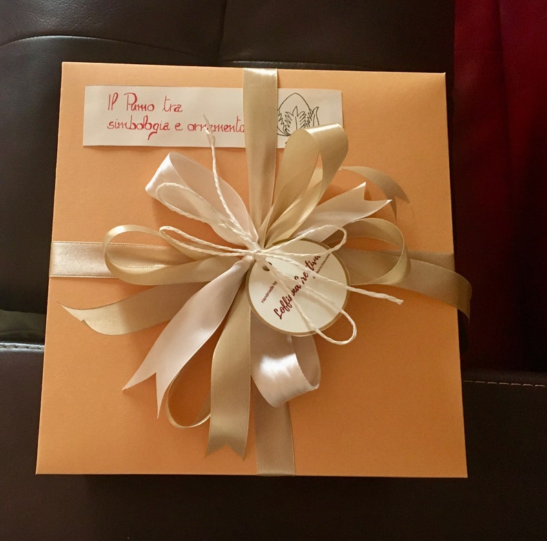 May include: A light orange gift box with a gold and white ribbon tied in a bow. The box has a handwritten label that says "Il Ramo tra simbologia e ornamento" and a smaller label that says "Loffy, un mare di fiori".