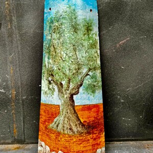 May include: Hand-painted tile featuring a green olive tree with a brown trunk and branches against a blue sky and orange ground. The tile is surrounded by white stones and has a signature in the bottom right corner.