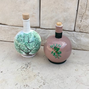 May include: Two ceramic olive oil bottles with cork stoppers. One bottle is white with a green olive tree painted on it. The other bottle is light pink with the word "OLIO" and a green olive branch painted on it.