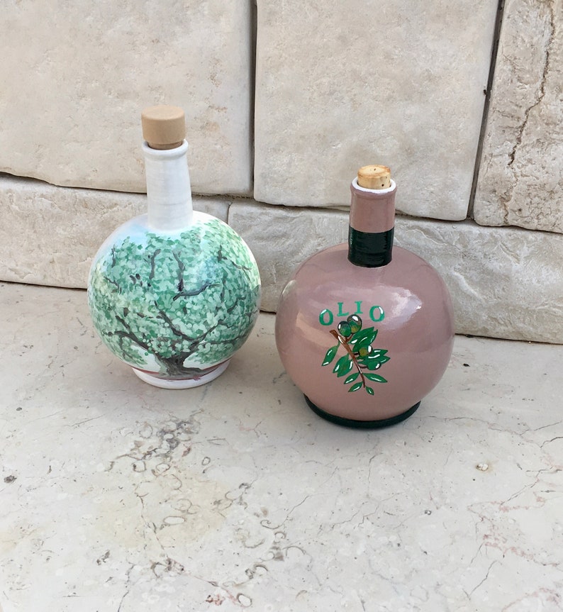 May include: Two ceramic olive oil bottles with cork stoppers. One bottle is white with a green olive tree painted on it. The other bottle is light pink with the word "OLIO" and a green olive branch painted on it.