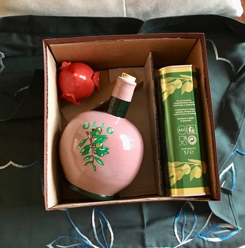 May include: A pink ceramic olive oil bottle with a green olive branch design and the word "OLIO" on the front. The bottle is in a brown cardboard box with a red ceramic pig figurine and a tin of olive oil.
