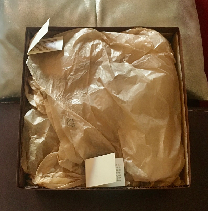 May include: A brown cardboard box with a brown paper tissue inside. The tissue is crumpled and has a small white tag on top.