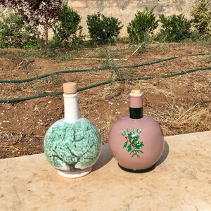 May include: Two ceramic olive oil bottles with cork stoppers. One bottle is white with a green olive tree design. The other bottle is pink with the word "OLIO" and a green olive branch design.