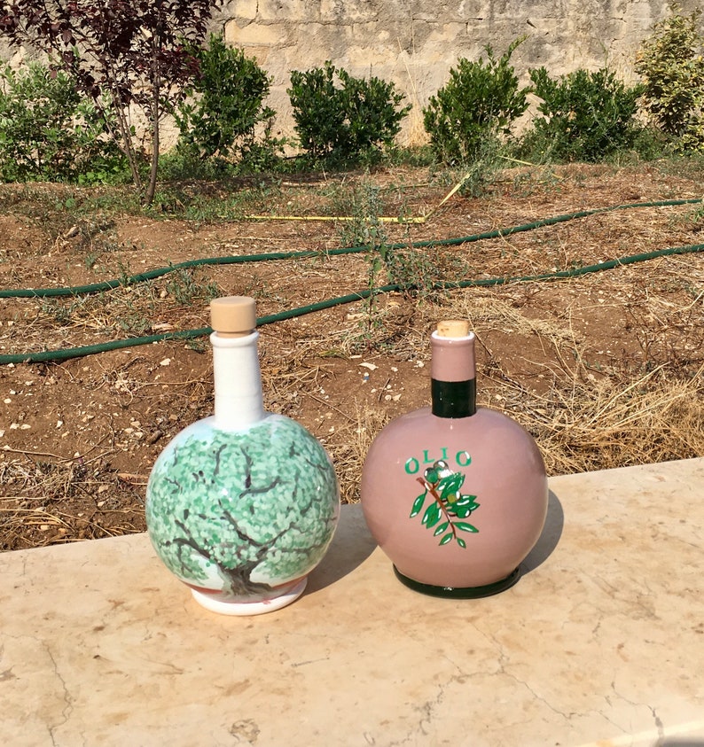 May include: Two ceramic olive oil bottles with cork stoppers. One bottle is white with a green olive tree design. The other bottle is pink with the word "OLIO" and a green olive branch design.