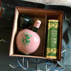 May include: A gift set featuring a pink ceramic olive oil bottle with a green olive branch design and the word "OLIO" on the front. The set also includes a green and gold tin with Italian text and a 1/2 liter content label.