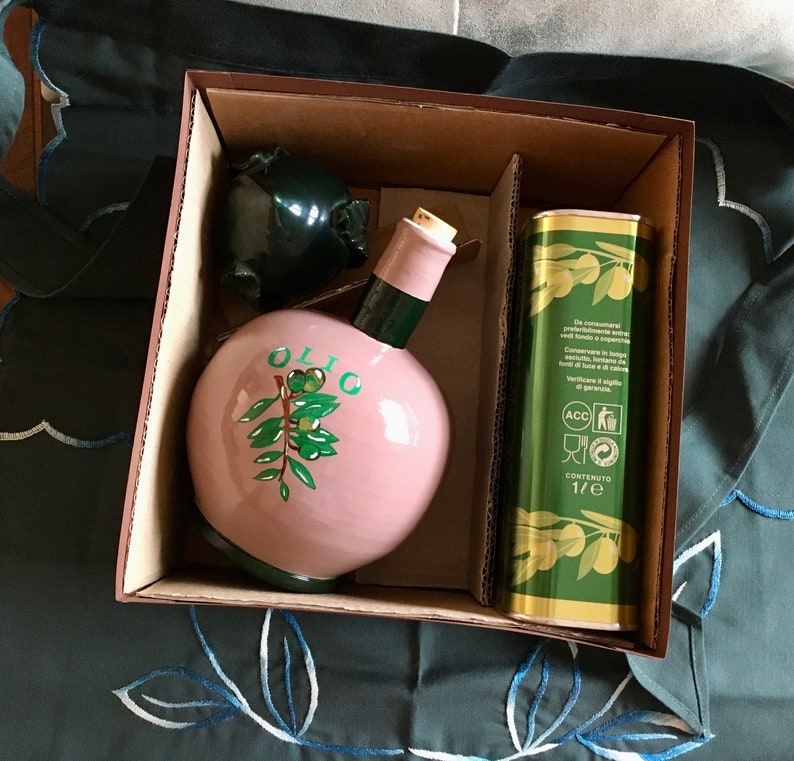 May include: A gift set featuring a pink ceramic olive oil bottle with a green olive branch design and the word "OLIO" on the front. The set also includes a green and gold tin with Italian text and a 1/2 liter content label.