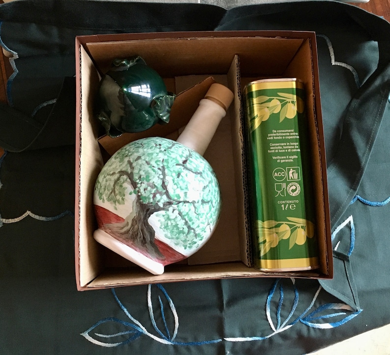 May include: A gift box set containing a ceramic olive oil bottle with a painted olive tree design, a green ceramic olive, and a bottle of olive oil. The olive oil bottle label reads "Contenuto 1/2 e".