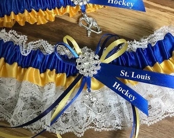May include: Two St. Louis Hockey garters. The top garter is blue and gold with a rhinestone embellishment and a hockey player charm. The bottom garter is white lace, blue and gold ribbon, and a rhinestone embellishment. The text "St. Louis Hockey" is on the blue ribbon.