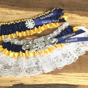 May include: A blue and gold garter with a rhinestone embellishment and two blue satin ribbons with the text "Michigan You're Next" and "Michigan Football".