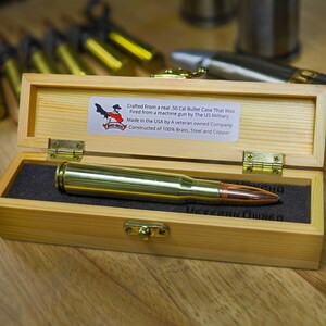 Peut inclure: Une boîte en bois contenant une douille de balle de calibre 50 en laiton. La boîte est ouverte, révélant la douille de balle. La boîte porte une étiquette indiquant "Crafted From a real 50 Cal Bullet Case That Was Fired from a machine gun by The US Military. Made in the USA by A veteran owned Company. Constructed of 100% Brass, Steel and Copper."