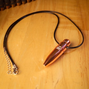 Puede incluir: Un collar con colgante en forma de bala de color cobre con un cordón negro. El colgante tiene líneas horizontales y un acabado brillante. El collar se muestra sobre una superficie de madera.