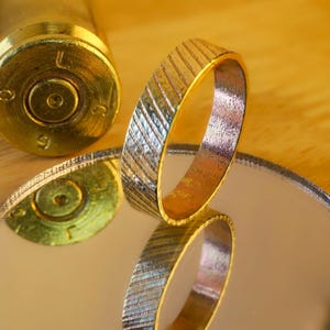 May include: A gold-colored ring with a textured, diagonal pattern. The ring is reflected in a mirrored surface. A brass bullet casing is in the background. The ring's design is detailed and eye-catching.