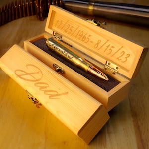 Pode incluir: Uma caixa de madeira com a palavra "Dad" gravada na tampa. No interior, uma caneta dourada com as palavras "A life well lived" é exibida. A caixa também tem datas gravadas: 12/25/1965-8/15/23.