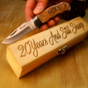 May include: A pocket knife with a wooden handle engraved with the name "MIKE" and a stainless steel blade, resting on a wooden box. The box is engraved with the text "20 Years And Still Sharp".