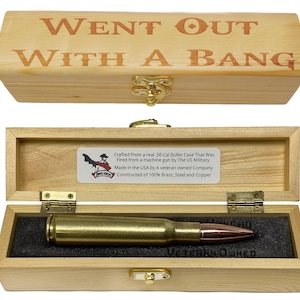 Puede incluir: Una caja de madera con el texto "WENT OUT WITH A BANG" grabado en la tapa. En el interior, un casquillo de bala .50 Cal descansa sobre un inserto de espuma negra. La caja también contiene una etiqueta con texto adicional.