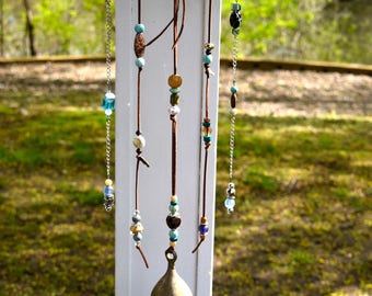 May include: A wooden branch with leather cords and metal chains hanging from it. The cords and chains are adorned with colorful beads, including blue, green, brown, and gold. A brass bell hangs from the bottom of the branch.