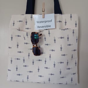 May include: Cream tote bag with a navy leaf pattern and navy handles. The bag has a front pocket and sunglasses hanging from it. A small card clipped to the bag reads "Waterproof Reversible."