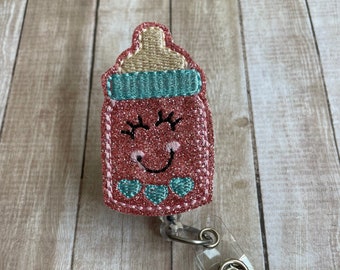 May include: Pink glitter baby bottle with a smiling face and teal accents. The bottle is attached to a clear plastic badge holder with a silver clip.