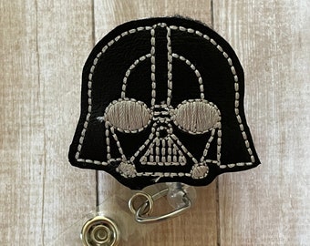 May include: Black embroidered badge reel featuring Darth Vader's helmet design. The design is stitched with silver thread on a black background. The badge reel has a clear plastic retractable cord and a metal clip.