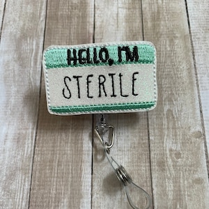 Op de afbeelding: Een wit en groen naambordje met de tekst "Hello, I'm Sterile". Het naambordje is bevestigd aan een transparante, intrekbare badgehouder van plastic.