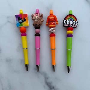 Puede incluir: Cuatro bolígrafos coloridos con adornos decorativos. Uno tiene un adorno cuadrado con las palabras "Be Kind" sobre un arcoíris. Otro tiene un adorno de vaca marrón. El tercero tiene un adorno de uniforme rojo y naranja. El último bolígrafo tiene un adorno de arcoíris con las palabras "Chaos Coordinator".