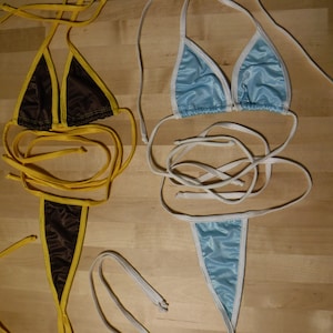 May include: Two bikinis on a wooden surface. One is brown with yellow trim and ties, the other is light blue with white trim and ties. Both have triangle tops and thong bottoms.