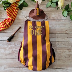May include: A brown and yellow striped baby onesie with the text "the chosen one" and a gold glitter number 1. A brown felt wizard hat with a gold glitter number 1 is also included.
