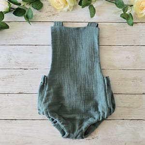 Könnte beinhalten: Ein salbeigrüner Baby-Strampler mit verstellbaren Trägern. Der Strampler ist aus einem weichen, leichten Stoff gefertigt und hat eine lockere Passform.
