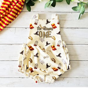 May include: Cream-colored baby romper with a repeating pattern of wizarding symbols, including hats, scarves, owls, and glasses. The text "the chosen ONE" is printed on the front. A red and gold striped fabric is visible on the left.
