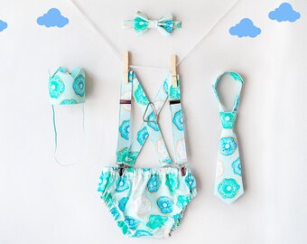 May include: A baby outfit set with a blue and white donut pattern. The set includes a crown, suspenders, and a bow tie.
