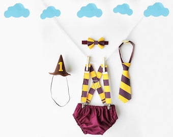 May include: A baby outfit for a first birthday party, featuring a brown witch hat with a yellow number one, a burgundy bow tie, burgundy and yellow striped braces, a burgundy and yellow striped tie, and burgundy bloomers.