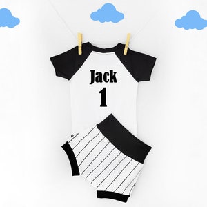 May include: A white and black baby outfit, including a short-sleeved shirt and matching shorts, hanging on a clothesline. The shirt has black sleeves and the name "Jack" and the number "1" printed on the front. The shorts have black and white stripes.