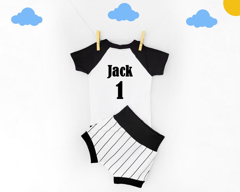 May include: A white and black baby outfit, including a short-sleeved shirt and matching shorts, hanging on a clothesline. The shirt has black sleeves and the name "Jack" and the number "1" printed on the front. The shorts have black and white stripes.