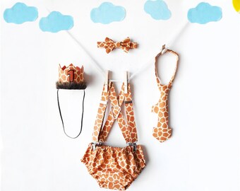 May include: A giraffe-print baby outfit for a first birthday celebration. The outfit includes a crown, suspenders, a bow tie, and a tie. The crown has a number one on it.
