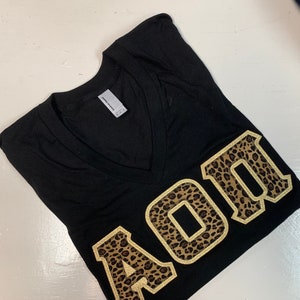 May include: Black v-neck t-shirt with a gold and brown leopard print "AOII" design.