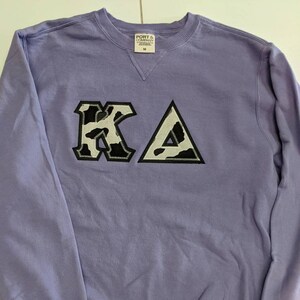 May include: A lavender sweatshirt with a black and white cow print "KA" embroidered on the front. The sweatshirt is a size medium and is made by Port & Company.