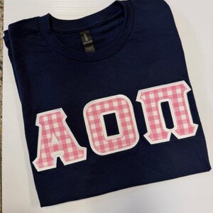 May include: Navy blue t-shirt with the letters A, O, and Π in a pink gingham pattern. The letters are outlined in white and sewn onto the shirt. The shirt is folded and lying on a white surface.