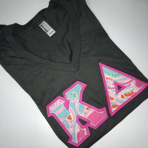 May include: A dark gray v-neck t-shirt with a pink and blue patterned "KA" applique.