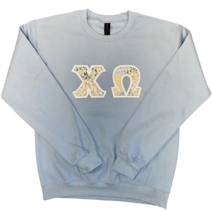 May include: A light blue sweatshirt with the Greek letters X and Ω appliqued on the front. The letters are white with a floral pattern in yellow, green, and white. The sweatshirt has long sleeves and a crew neck.