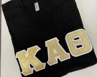 Camisa con cuello en V y letras Kappa Alpha Theta en negro con letras griegas en dorado metalizado y blanco