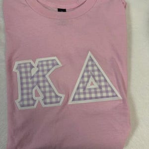 May include: A light pink t-shirt with the letters "KA" and a triangle, all in a white and lavender gingham pattern. The shirt is folded and appears to be made of soft material.
