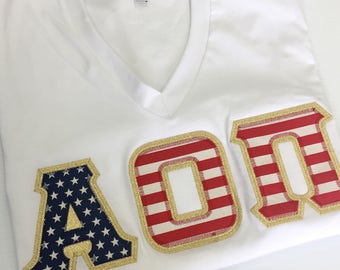 Camisa de cuello en V con letras de la hermandad griega en blanco con letras de la bandera estadounidense