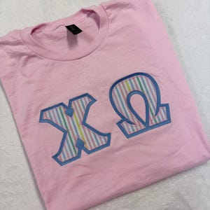 May include: A light pink t-shirt with the Greek letters X and Ω in a blue outline. The letters are filled with pastel rainbow stripes. The shirt is folded on a white surface.