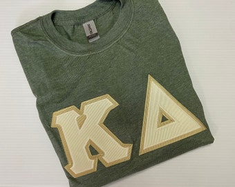 May include: A green t-shirt with a white and gold "KA" embroidered on the front.