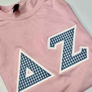 May include: A pink t-shirt with a blue and white gingham "AZ" design. The letters are outlined in white.