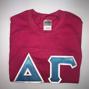 May include: A pink t-shirt with a blue and white embroidered Greek letter design that says "A". The shirt is folded and the Gildan Heavy Cotton tag is visible.