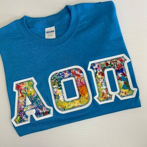 May include: A blue t-shirt with a white outline of the letters "AOII" in a colorful, paint-splattered design.