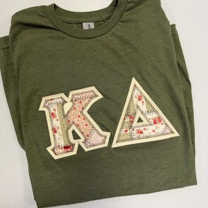 May include: Olive green t-shirt with the letters "KA" appliqued on the front. The letters are cream-colored with floral fabric inserts. The "K" is a block letter, and the "A" is a triangle. The shirt is made of a soft, heathered fabric.
